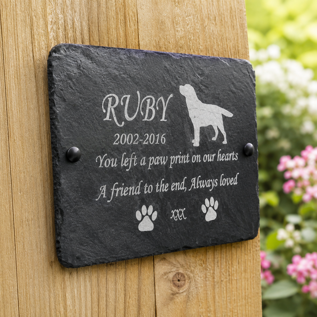 Personalized black slate memorial plaque for a dog named RUBY, mounted on a wooden fence with a blurred garden background. The plaque features engraved white text, paw prints, and a dog silhouette.