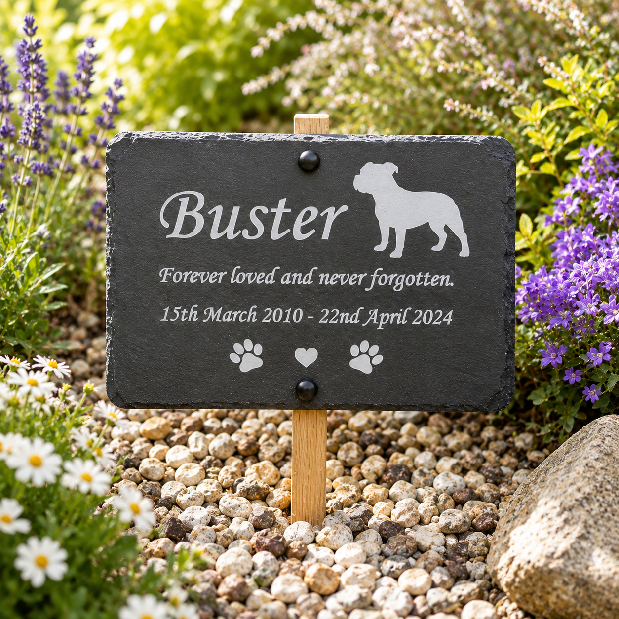 Personalized black slate pet memorial garden sign with engraved white text and silhouette of a bulldog, mounted on a wooden stake in a garden bed with gravel, flowers, and greenery.