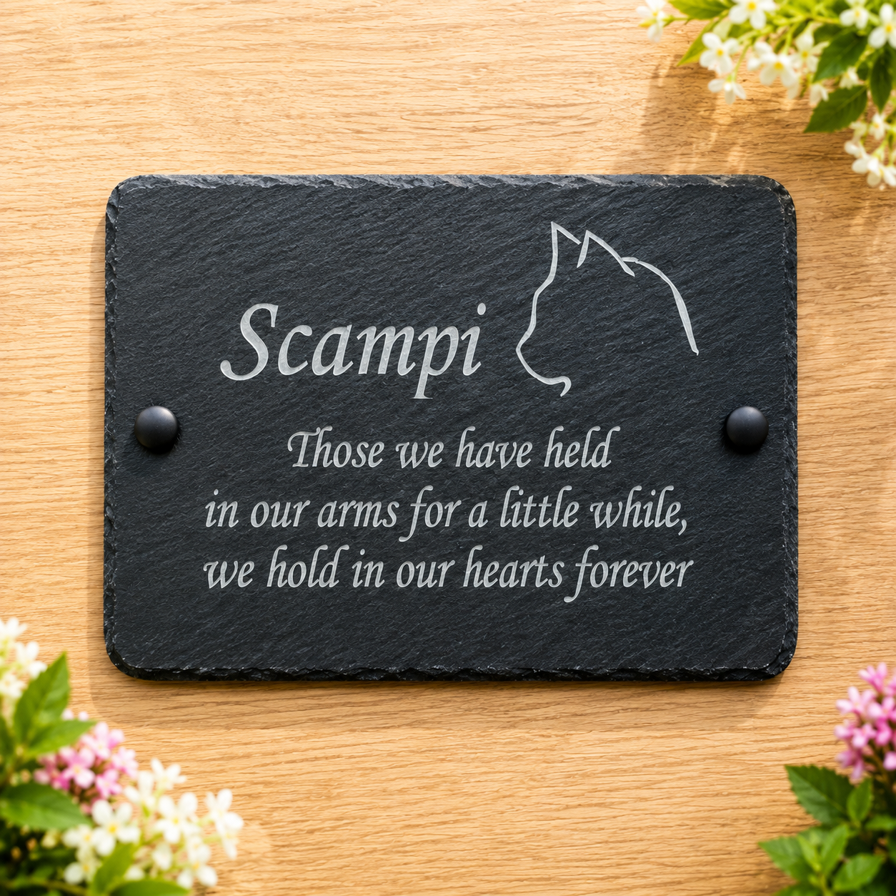 A rectangular black slate memorial plaque with engraved white text and a cat silhouette, displayed on a light wood surface with small flowers around the edges. The plaque reads: 'Scampi Those we have held in our arms for a little while, we hold in our hearts forever.