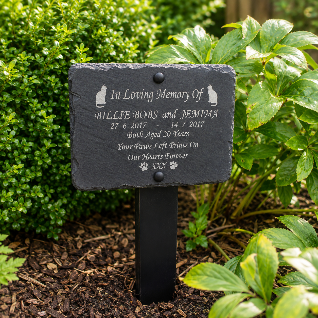 A personalized black slate pet memorial garden plaque mounted on a black metal stake, set in a garden bed with green foliage and mulch.