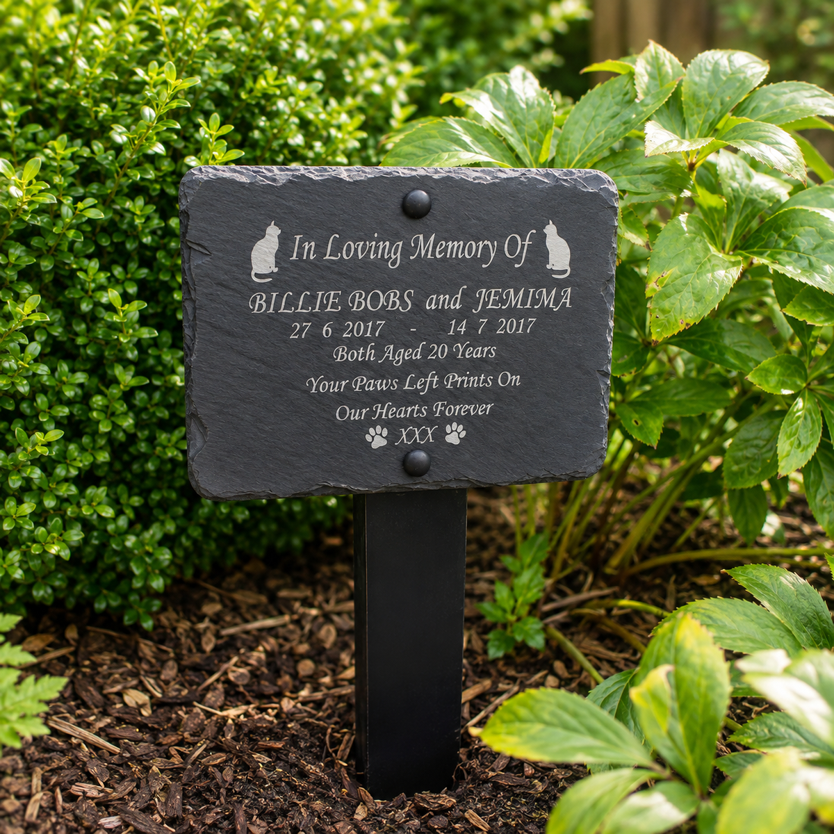 A personalized black slate pet memorial garden plaque mounted on a black metal stake, set in a garden bed with green foliage and mulch.
