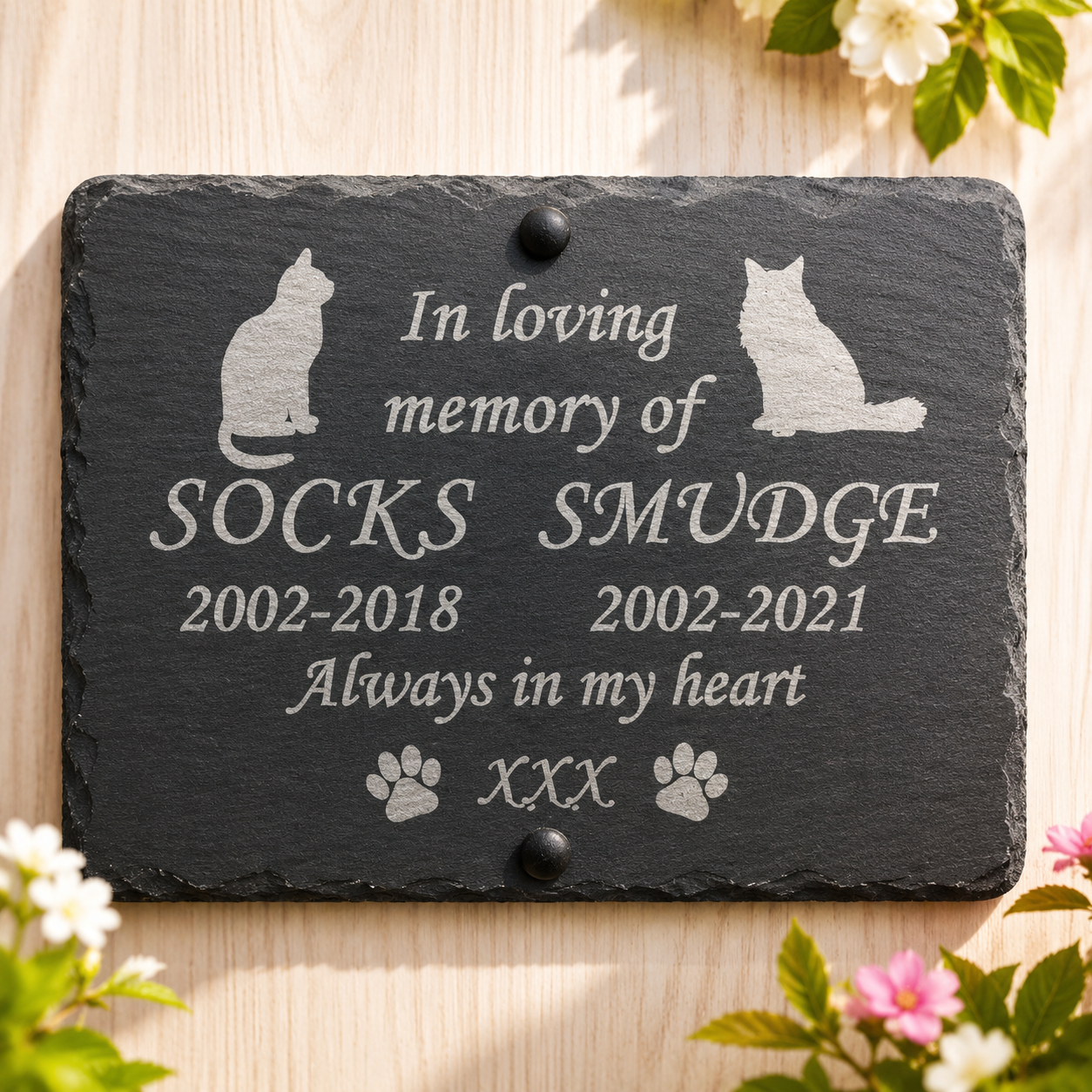 A rectangular dark slate memorial plaque with engraved silver text reading 'In loving memory of SOCKS SMUDGE 2002-2018 2002-2021 Always in my heart' with cat and paw print illustrations, photographed on a light wood surface with flowers around the edges.
