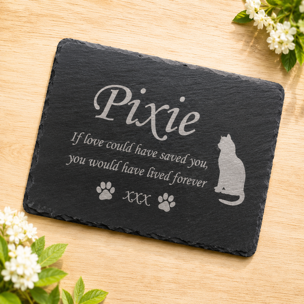 Personalized black slate plaque for a pet, engraved with the name 'Pixie', a cat silhouette, paw prints, and the phrase 'If love could have saved you, you would have lived forever'. The plaque is displayed on a light wood surface with decorative white flowers in the corners.