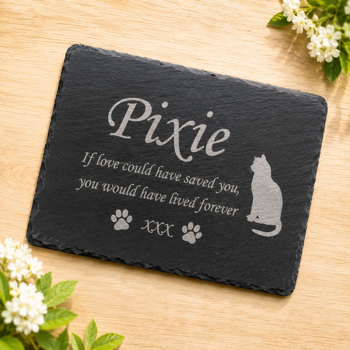 Personalized black slate plaque for a pet, engraved with the name 'Pixie', a cat silhouette, paw prints, and the phrase 'If love could have saved you, you would have lived forever'. The plaque is displayed on a light wood surface with decorative white flowers in the corners.