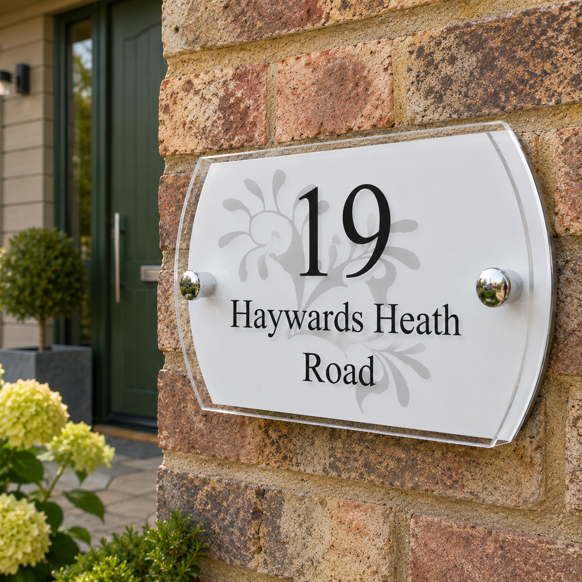 A modern white house number plaque mounted on a brick wall, displaying '19 Haywards Heath Road' with a subtle floral design behind the text.