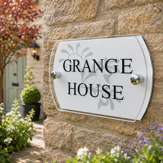 A white arched house sign with black lettering reading 'GRANGE HOUSE' and a subtle gray floral motif, mounted on a beige stone wall with garden plants visible around it.