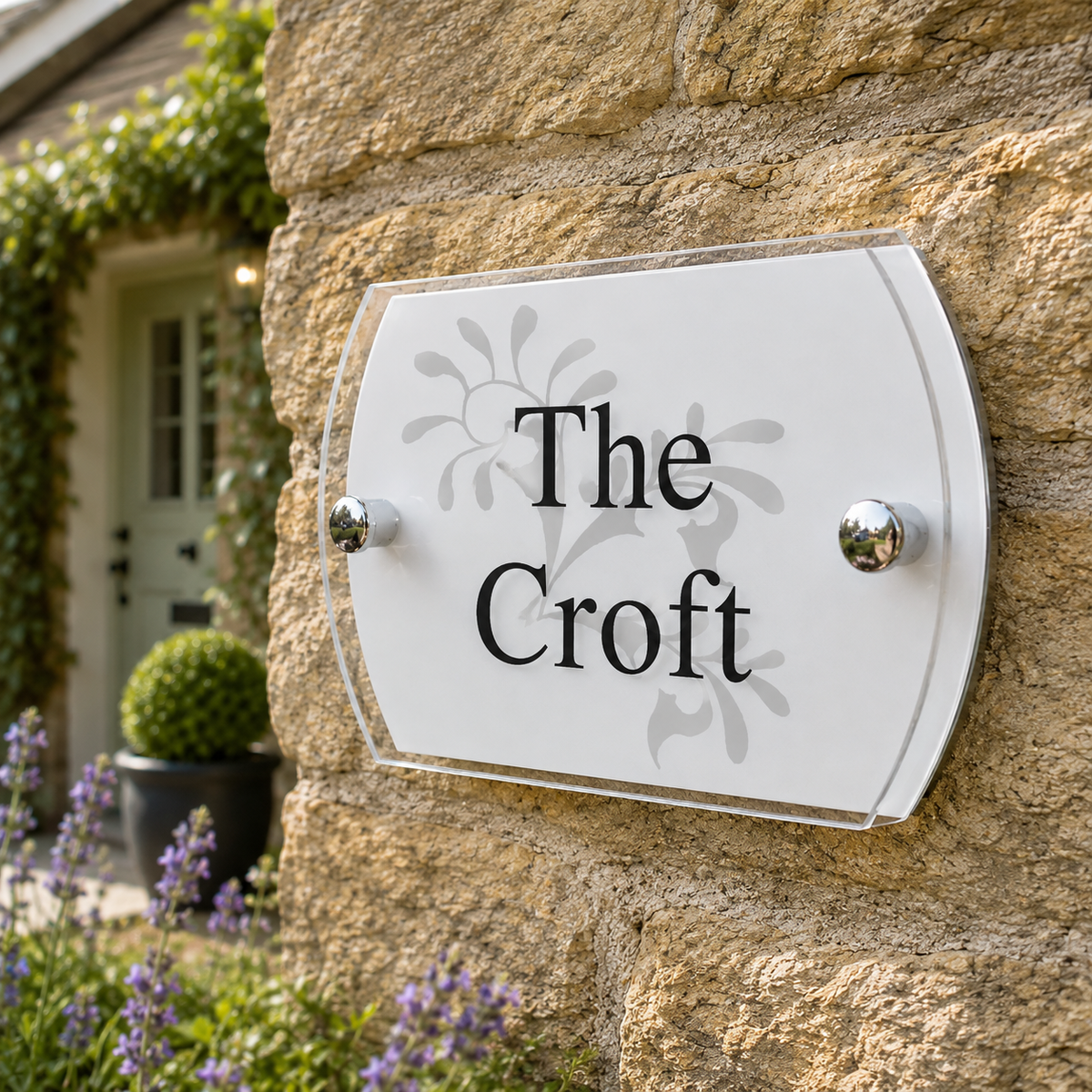 A custom white acrylic house plaque with black lettering reading 'The Croft' and a subtle floral design, mounted on a rustic stone wall with garden plants in the background.