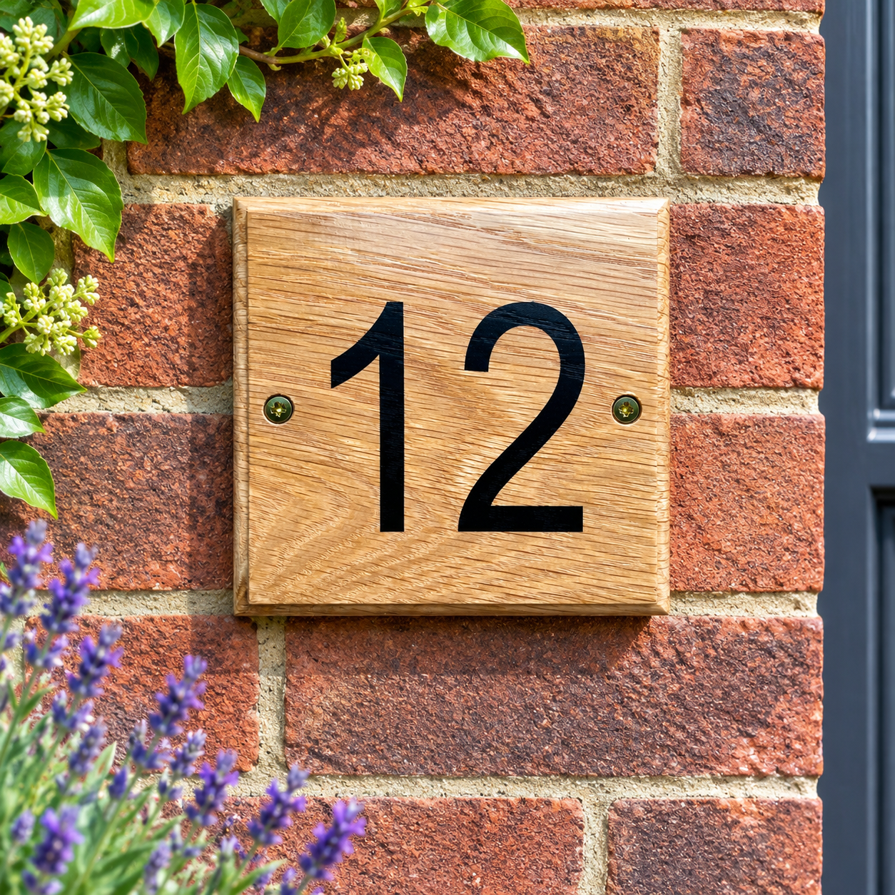 A square wooden house number plaque displaying '12' in black, mounted on a red brick wall with green leaves and purple flowers in the foreground.