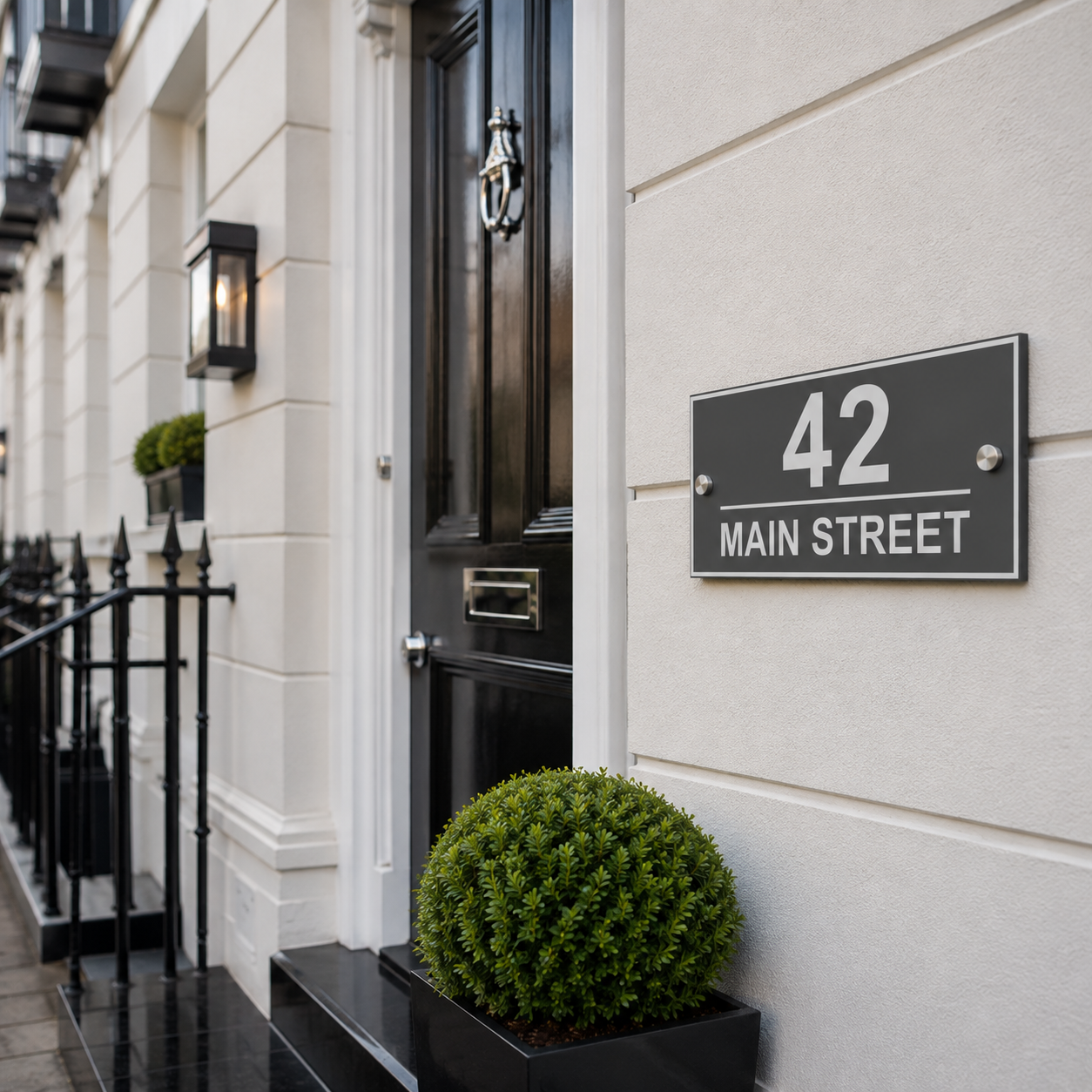 A black house number 42 sign reading 'MAIN STREET' mounted on a cream-colored stone wall next to a black front door and a small green shrub.