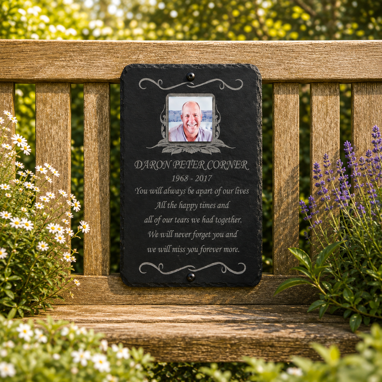 A personalized black slate memorial plaque hanging on a wooden garden bench, featuring a photo of a man and engraved text reading 'DARON BETER CORNER 1968-2017' with a remembrance poem. The background shows a lush green garden with white and purple flowers.