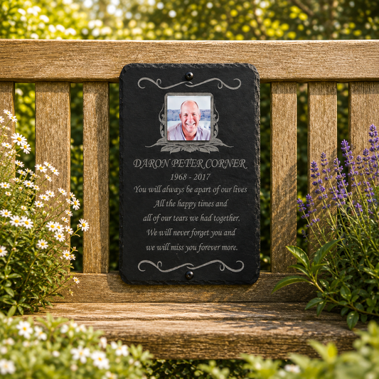 A personalized black slate memorial plaque hanging on a wooden garden bench, featuring a photo of a man and engraved text reading 'DARON BETER CORNER 1968-2017' with a remembrance poem. The background shows a lush green garden with white and purple flowers.