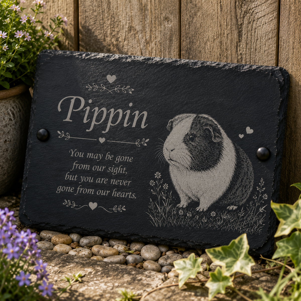 Personalized black slate memorial plaque with engraved text reading 'Pippin' and a detailed guinea pig illustration, displayed outdoors against a wooden fence with flowers and stones around it.
