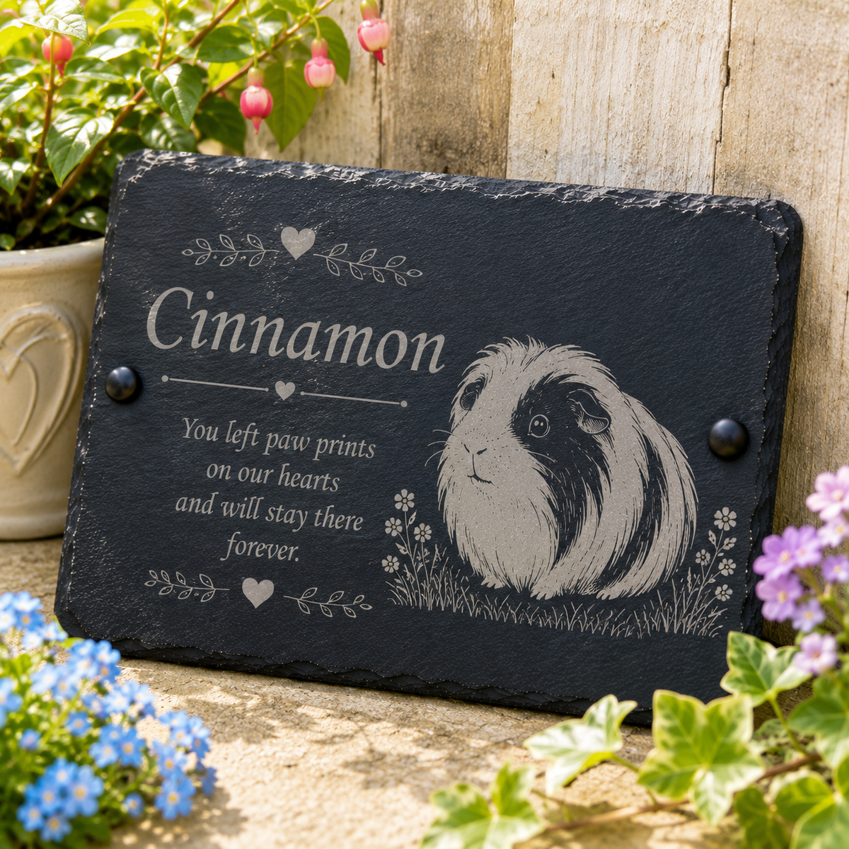 Personalized black slate memorial plaque with an engraved guinea pig illustration and the name 'Cinnamon,' displayed outdoors on a wooden surface with flowers and plants around it.
