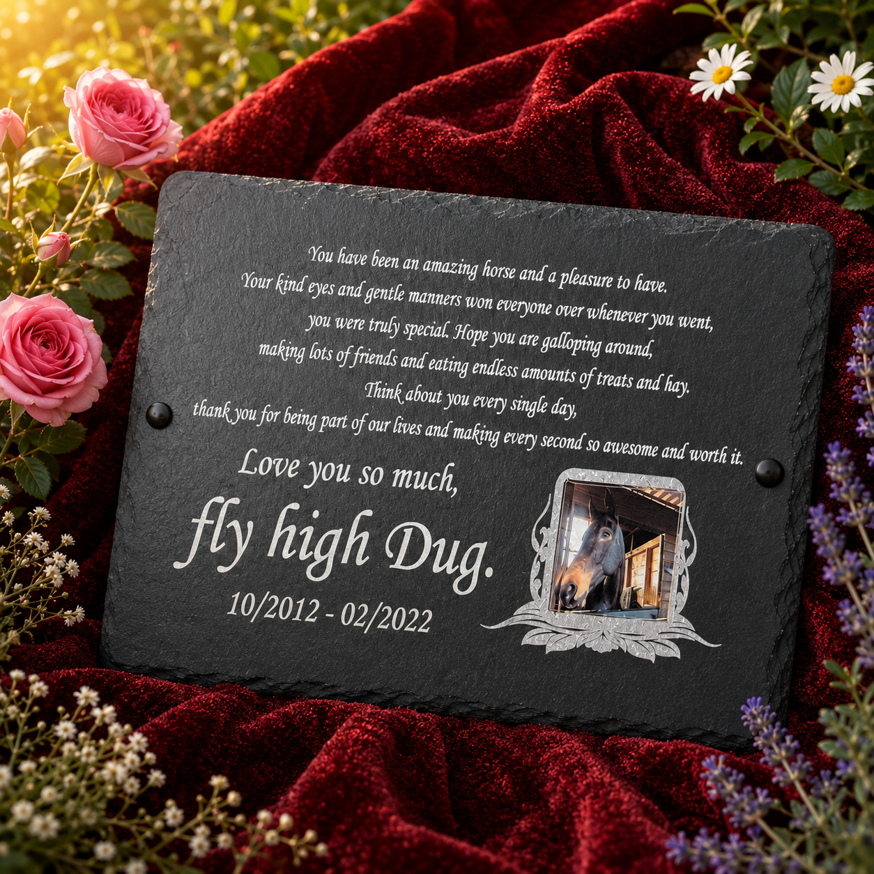 Personalized black slate memorial plaque with engraved white text and a small photo frame, displayed on a red velvet fabric with roses and daisies around it.