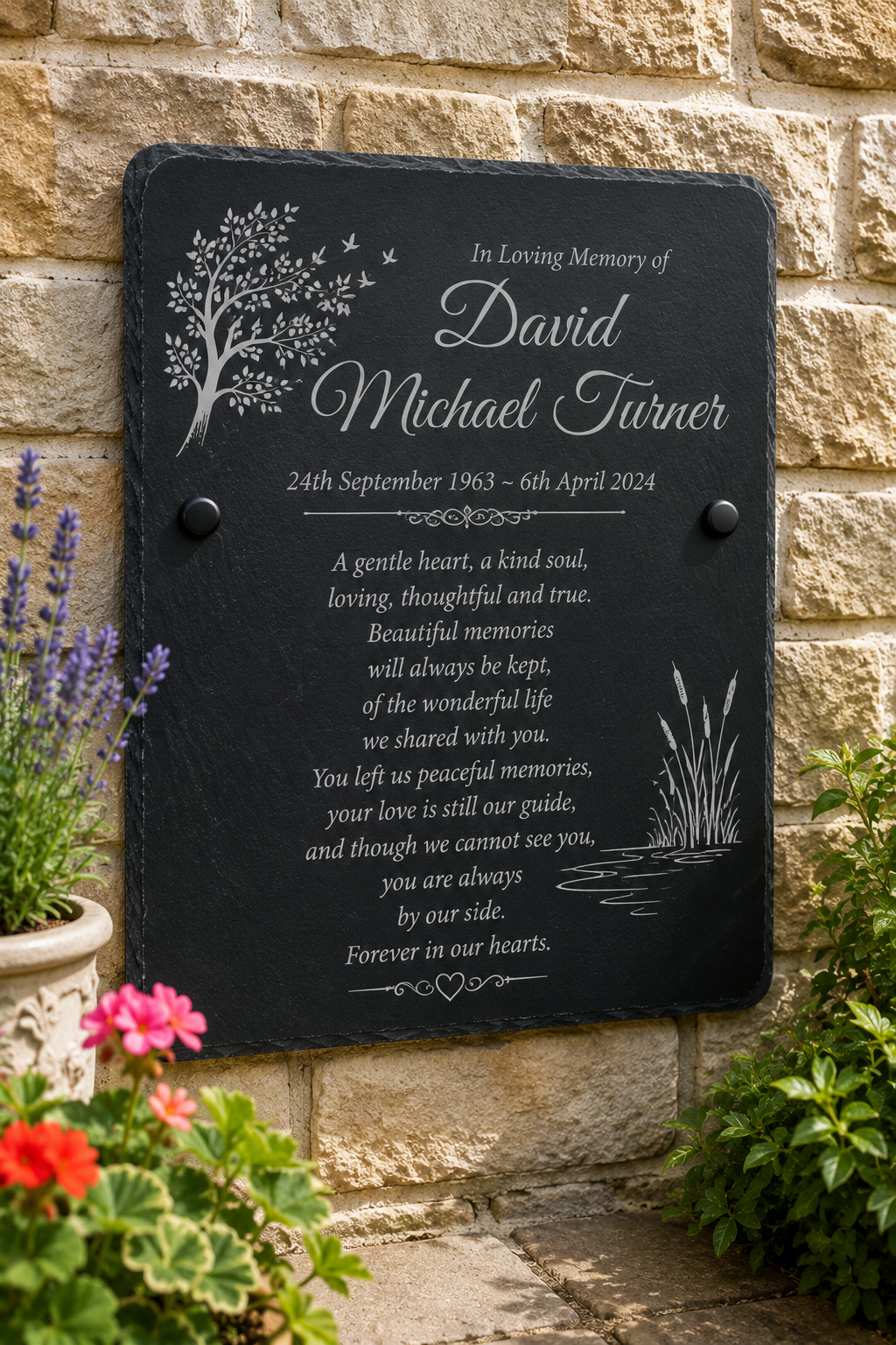 Personalized black slate memorial plaque with engraved white text and tree/water illustrations, mounted on a beige stone wall with flowers at the base.