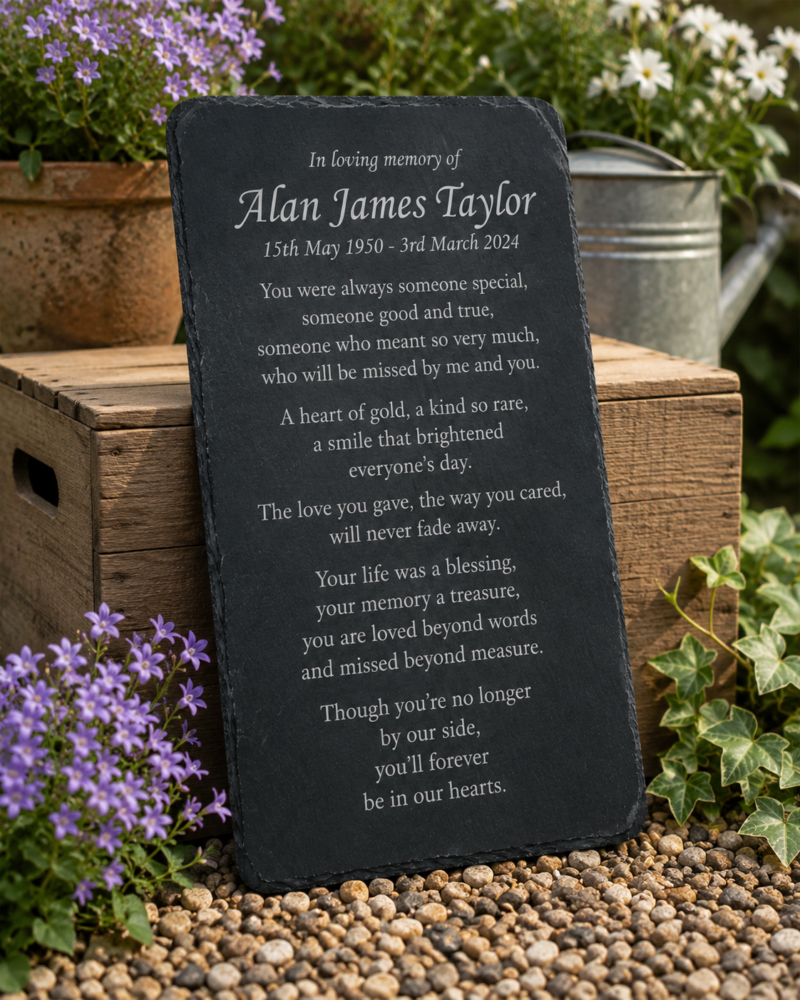 A personalized black slate memorial garden plaque with white engraved text, displayed outdoors on a bed of small pebbles next to a wooden crate and green plants.