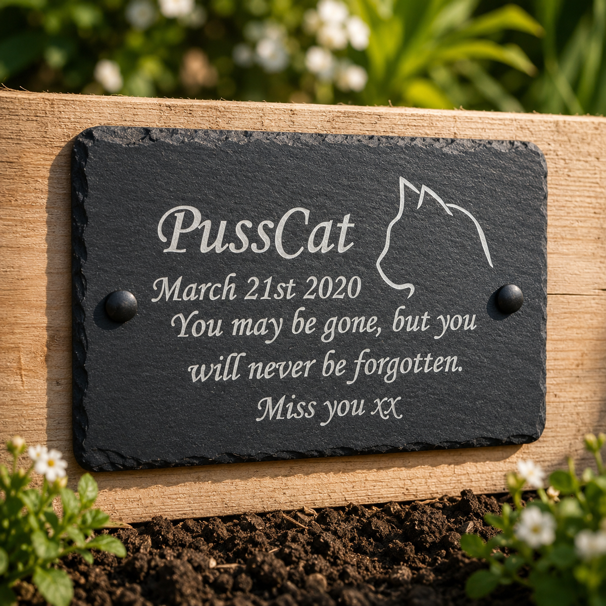 A personalized black slate pet memorial plaque with white engraved text and a cat silhouette, mounted on a wooden garden wall in soil with blurred greenery in the background.