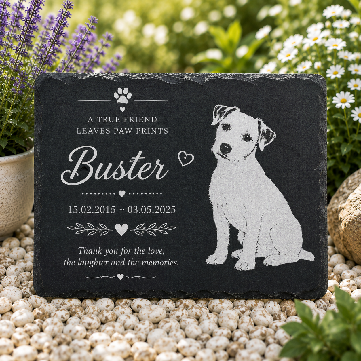 Personalized black slate pet memorial garden stone with engraved text and a white dog illustration, displayed on white pebbles outdoors with flowers in the background.