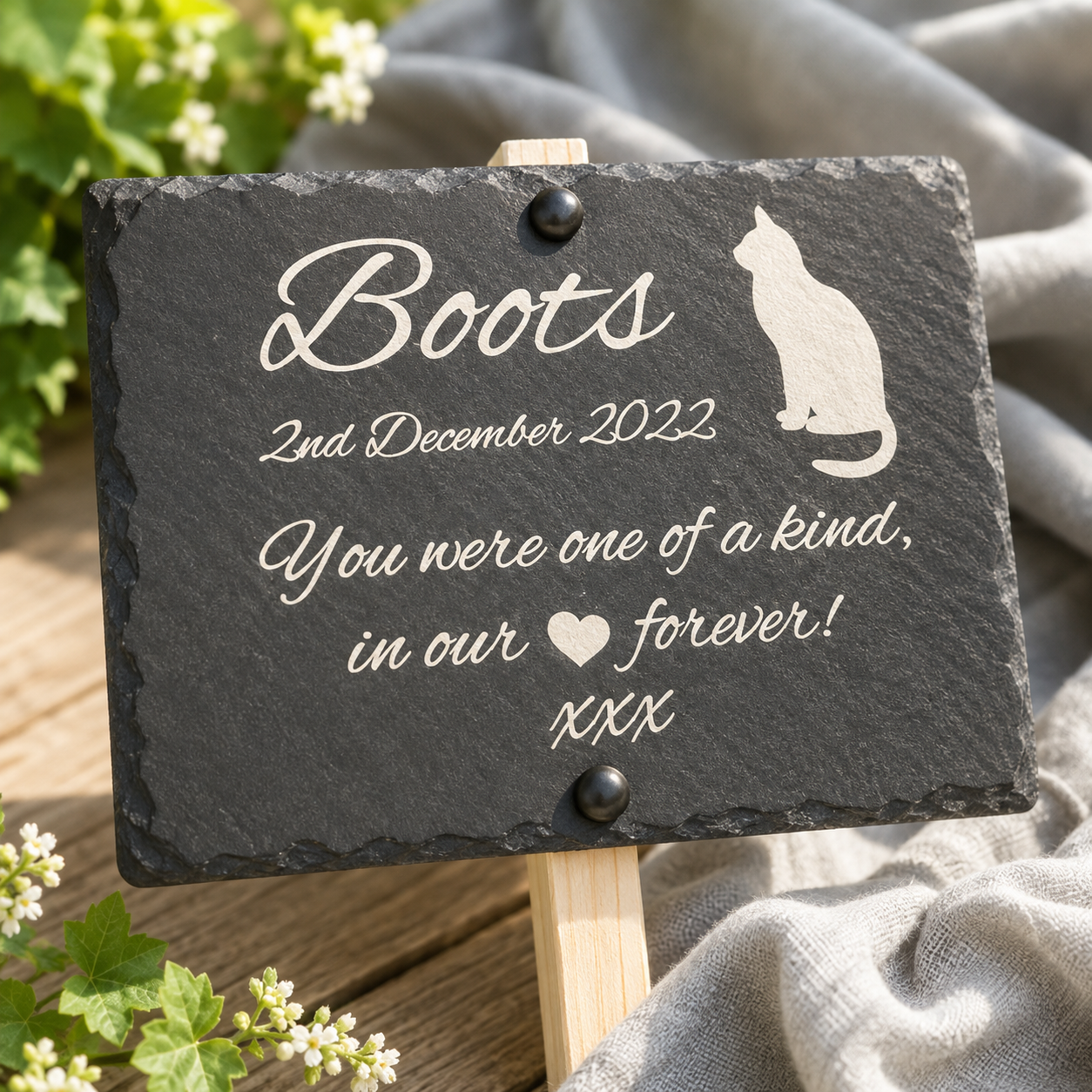 Personalized black slate memorial sign on a small wooden stand, engraved with the name 'Boots', a cat silhouette, and remembrance text, styled outdoors with greenery and draped fabric.
