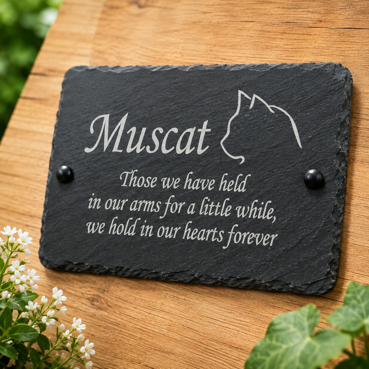 Personalized black slate memorial plaque with engraved white text reading 'Muscat' and a cat silhouette, mounted on a wooden surface with small black round studs. White flowers and green leaves are visible around the edges.