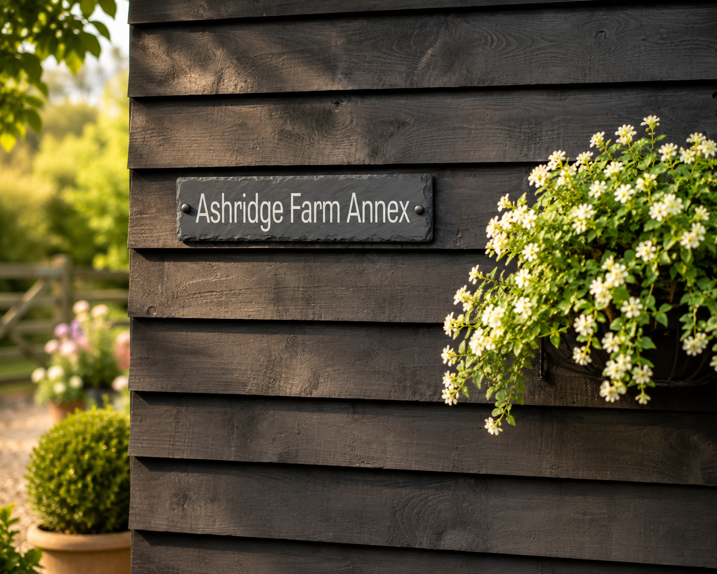 A dark slate sign reading 'Ashridge Farm Annex' mounted on a dark wood-paneled wall, with white flowering plants hanging from a basket on the right.