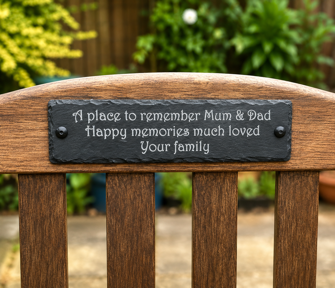 A personalized black slate memorial plaque mounted on the backrest of a wooden garden bench, with a blurred outdoor garden background.