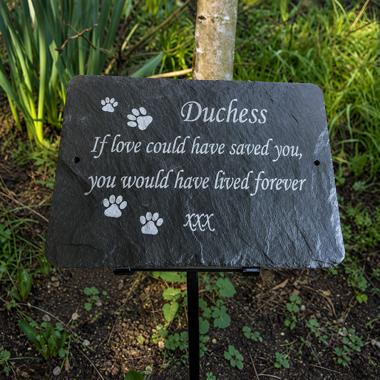 A personalized black slate pet memorial garden plaque with white engraved text and paw prints, mounted on a black metal stake in an outdoor garden setting.