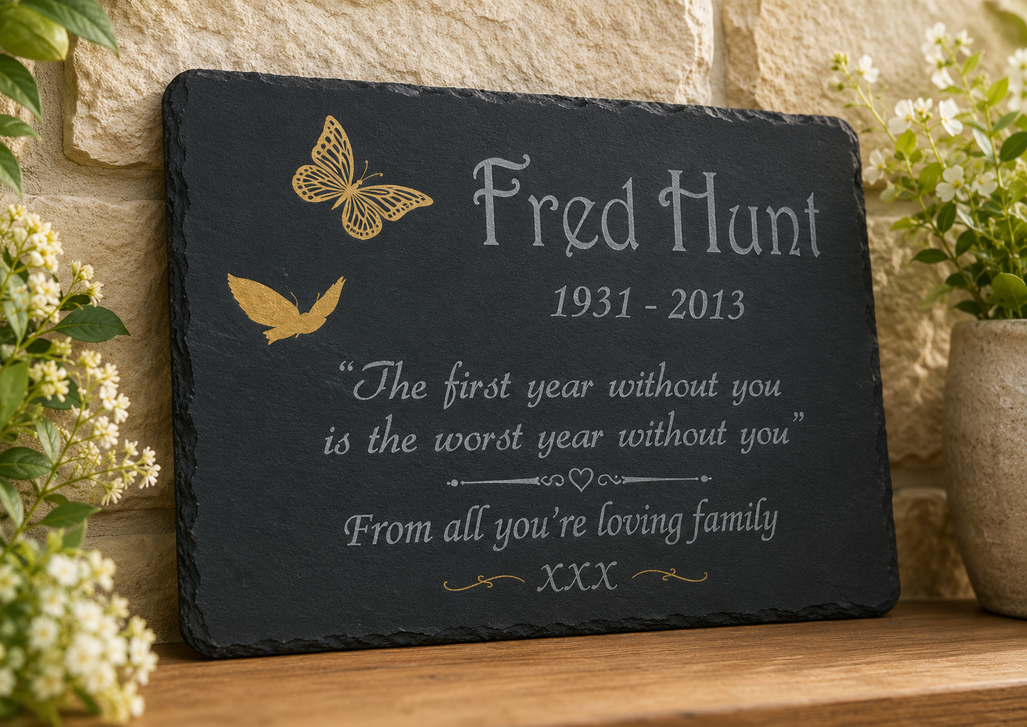 Personalized black slate memorial plaque with engraved white text reading 'Fred Hunt 1931-2013' and a remembrance quote, decorated with gold butterfly and bird illustrations, displayed on a wooden surface against a stone wall with flowers.