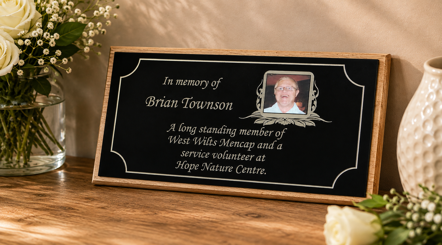 A personalized wooden memorial plaque with a black face and white text, featuring a photo of the deceased and engraved decorative borders. The plaque is displayed on a wooden table next to white roses in a glass vase and a white ceramic vase.