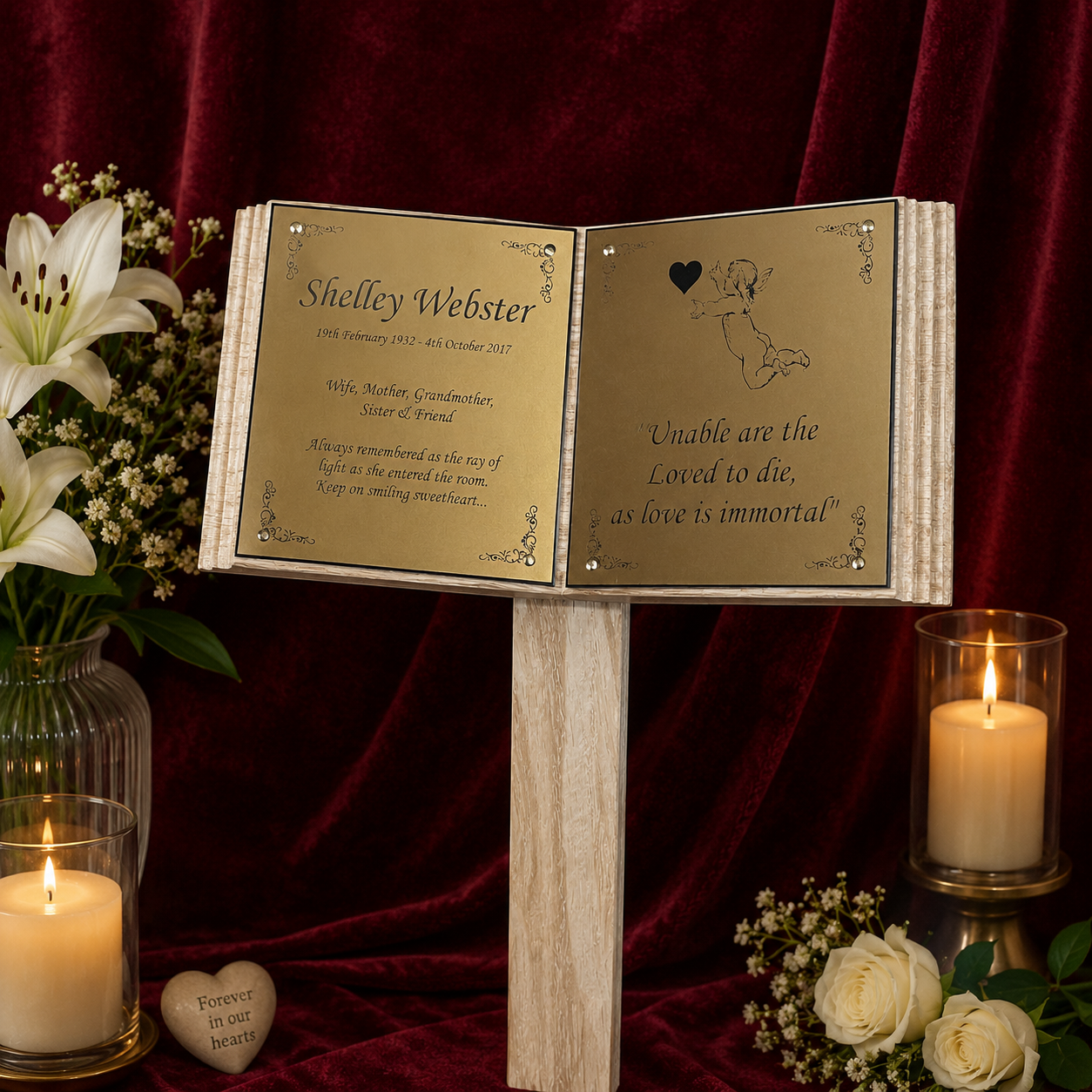 An open wooden keepsake book with gold-toned pages stands on a wooden block against a dark red velvet backdrop. The left page is personalized with the name Shelley Webster and a poem, and the right page has a small cherub illustration and the quote 'Unable are the Loved to die, as love is immortal.