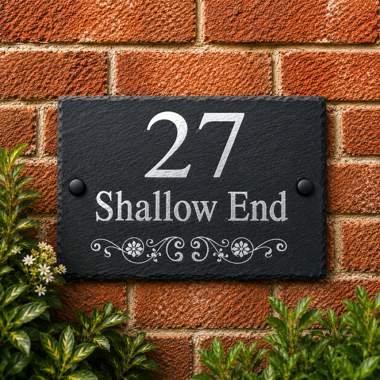 A rectangular black slate house number plaque mounted on a red brick wall, with silver lettering reading '27 Shallow End' and decorative scrollwork. Green foliage is visible at the bottom corners.
