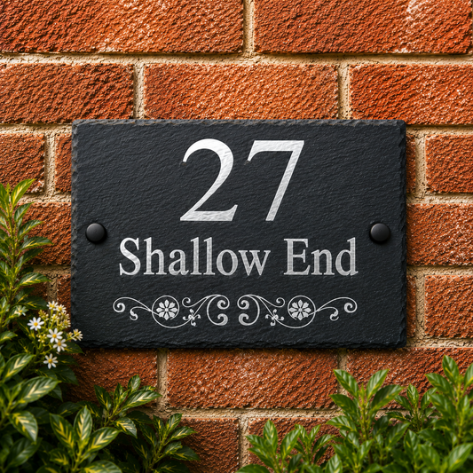 A rectangular black slate house number plaque mounted on a red brick wall, with silver lettering reading '27 Shallow End' and decorative scrollwork. Green foliage is visible at the bottom corners.