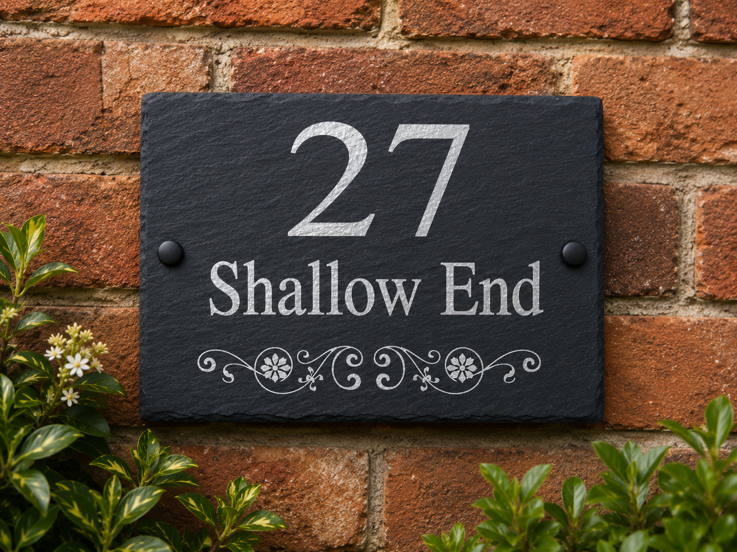 A rectangular black slate house number plaque mounted on a red brick wall, with white engraved text reading '27 Shallow End' and decorative scrollwork below, framed by green foliage at the bottom.