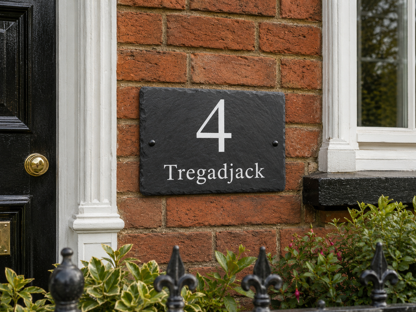 A rectangular black slate house number plaque reading '4 Tregadjack' mounted on a red brick wall next to a black front door and white window trim.