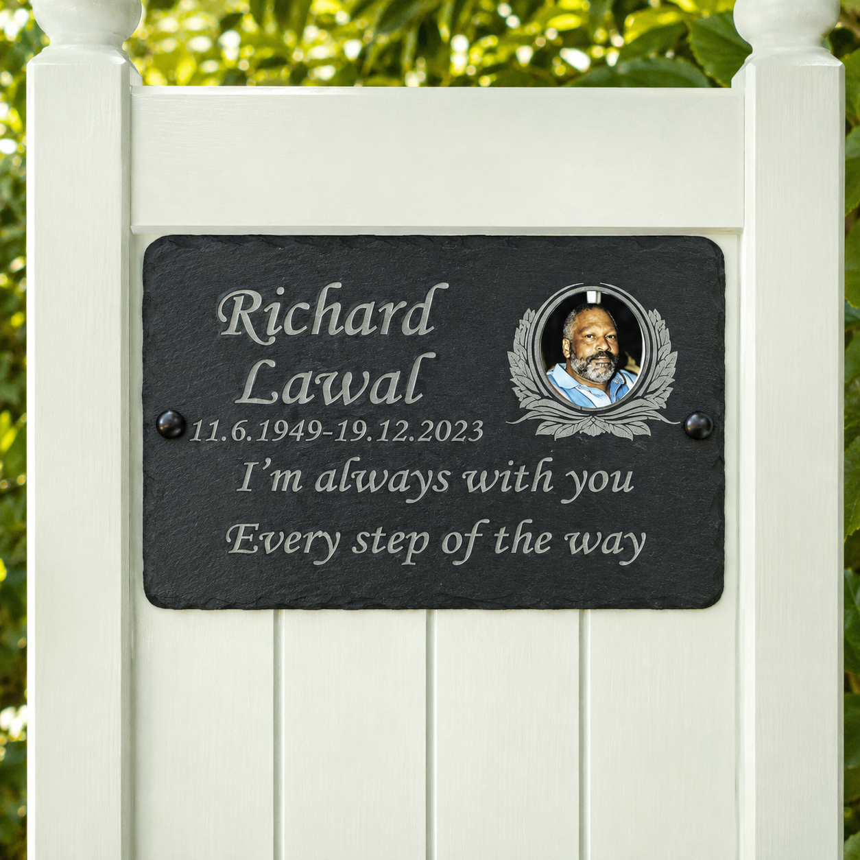 Personalized black slate memorial plaque mounted on a white wooden garden fence, featuring engraved silver text reading 'Richard Lawal' with birth and death dates and the phrase 'I’m always with you Every step of the way', plus a circular portrait photo.