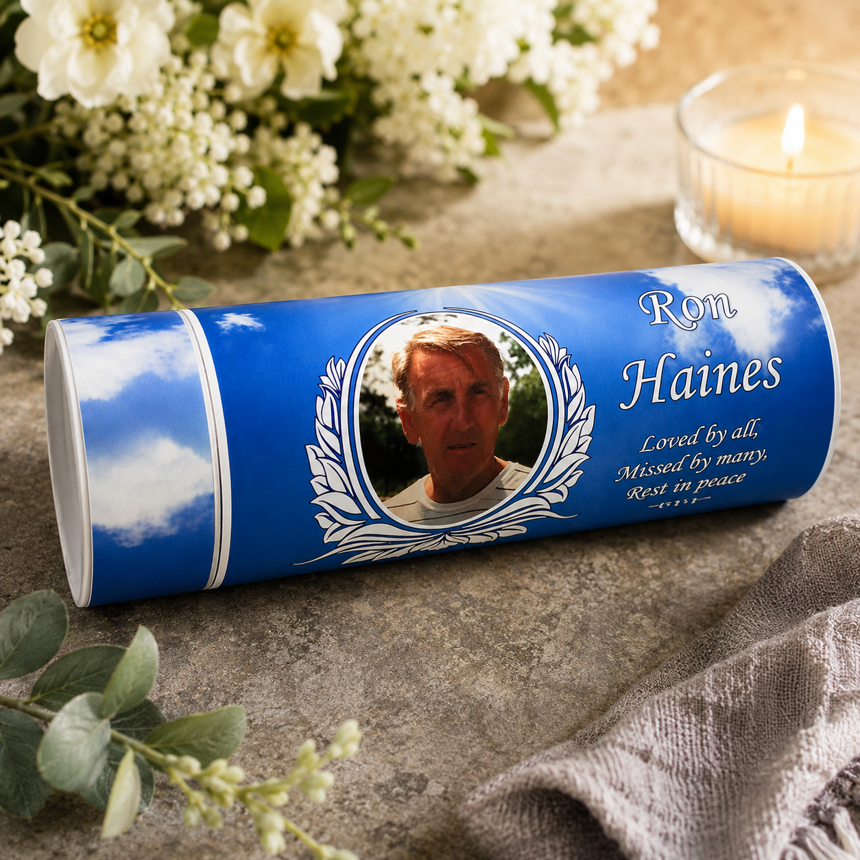 Personalized blue urn cylinder with a portrait of Ron Haines and memorial text, styled on a stone surface with flowers, greenery, and a lit candle in the background.