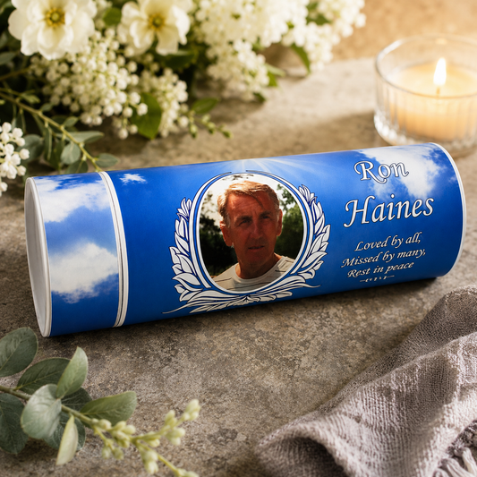 Personalized blue urn cylinder with a portrait of Ron Haines and memorial text, styled on a stone surface with flowers, greenery, and a lit candle in the background.