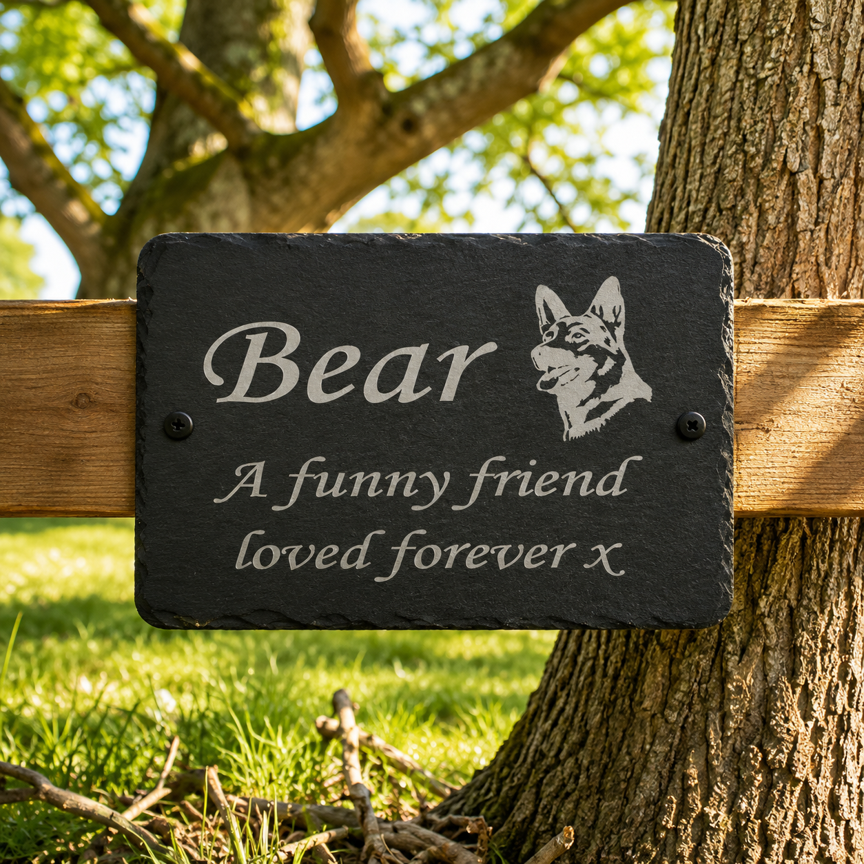 A rectangular black slate memorial garden plaque mounted on a wooden fence next to a tree trunk. The plaque is engraved with the word 'Bear', a dog portrait, and the text 'A funny friend loved forever x'.