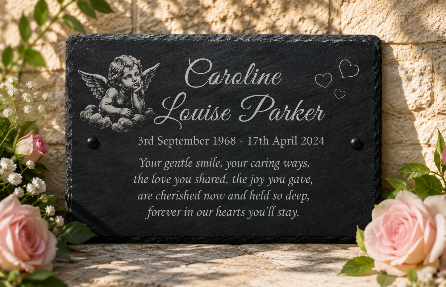 Personalized black slate memorial plaque with engraved cherub artwork and white text, displayed outdoors on a stone surface with pink roses and greenery around it.