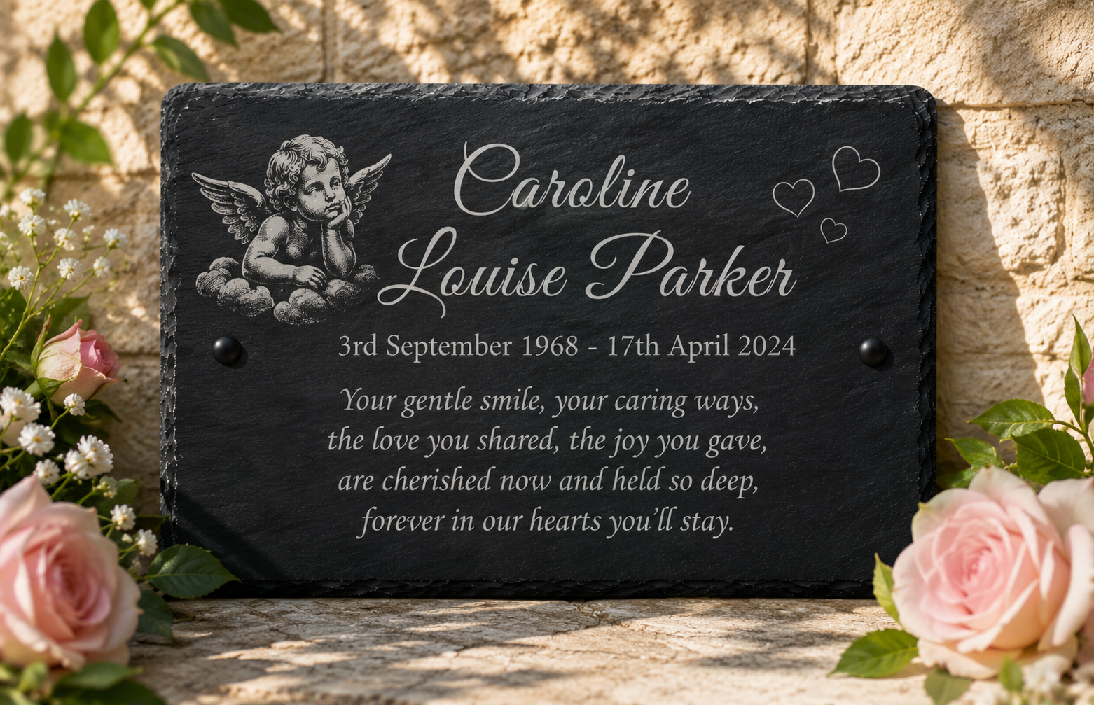 Personalized black slate memorial plaque with engraved cherub artwork and white text, displayed outdoors on a stone surface with pink roses and greenery around it.