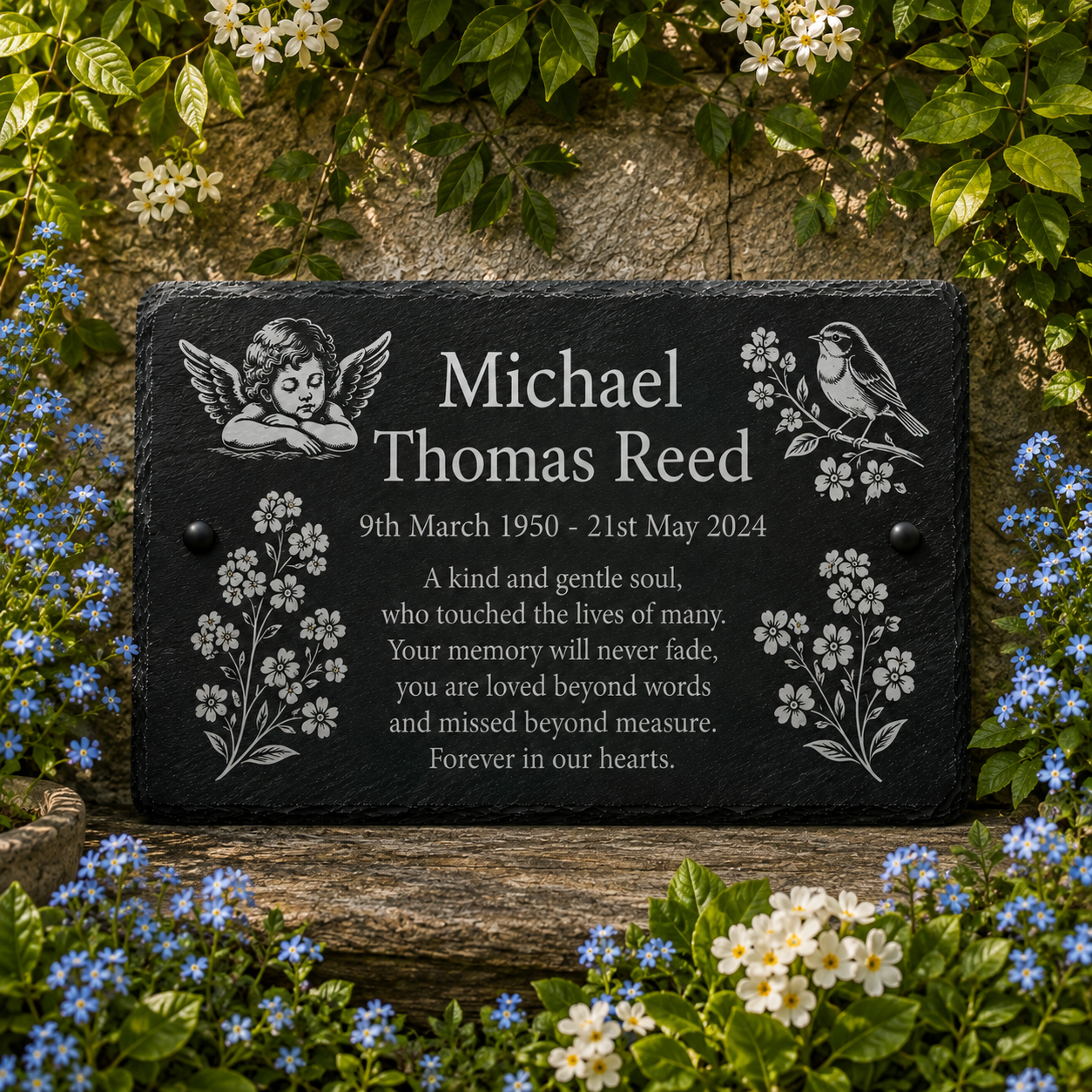Personalized black slate memorial plaque with engraved cherub, bird, flowers, and memorial text, displayed outdoors on a wooden surface surrounded by green leaves and blue/white flowers.