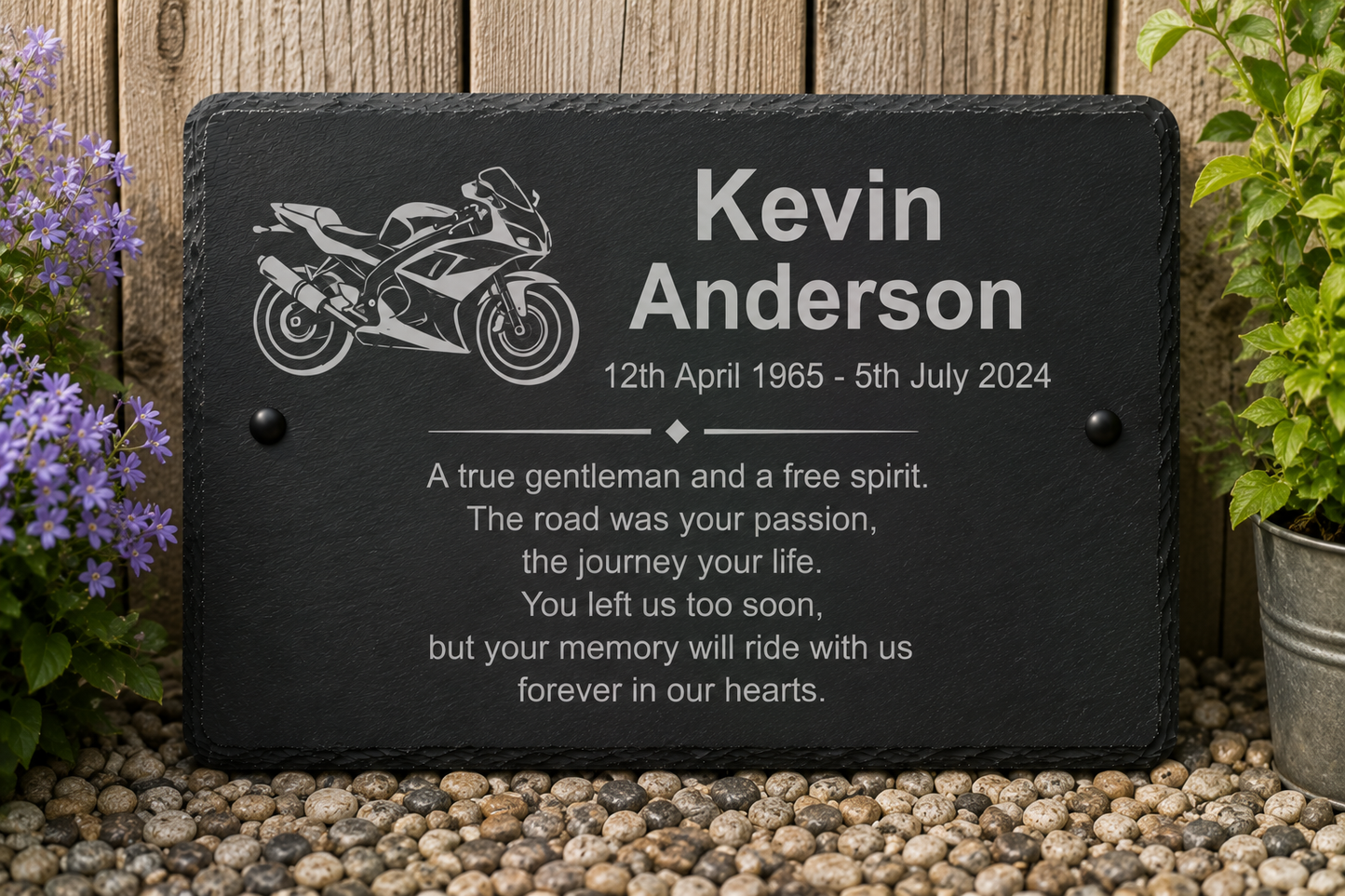 Personalized black slate memorial plaque with an engraved motorcycle illustration and the name Kevin Anderson, placed outdoors on pebbles against a wooden fence with purple flowers and greenery.