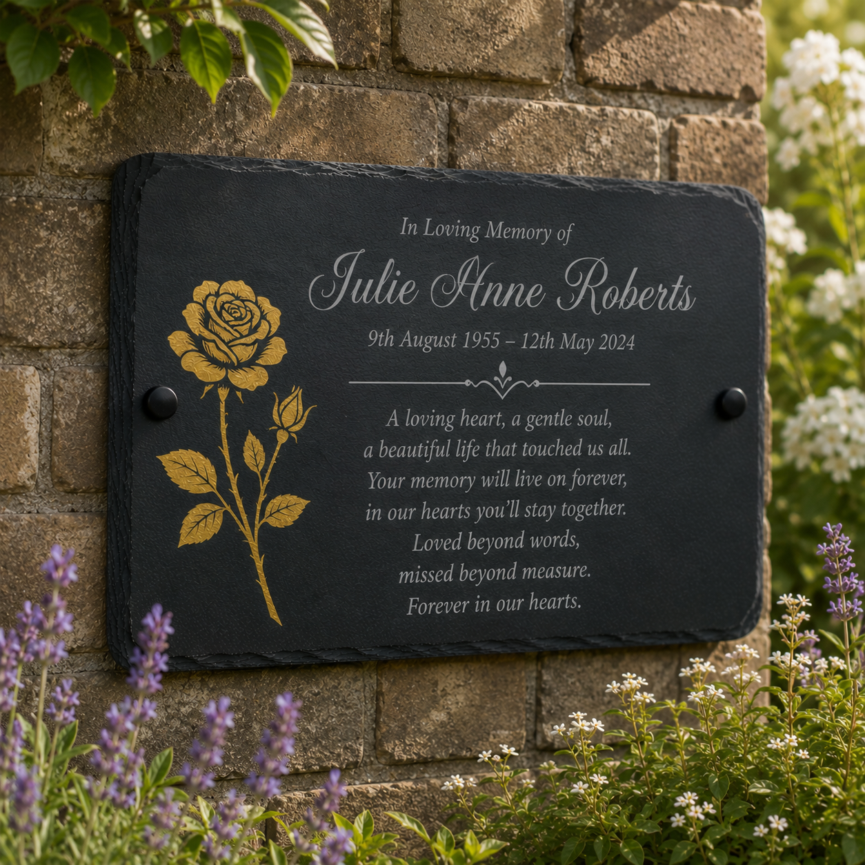 A personalized black slate memorial plaque with a gold rose engraving, mounted on a brick wall with garden plants in the foreground.