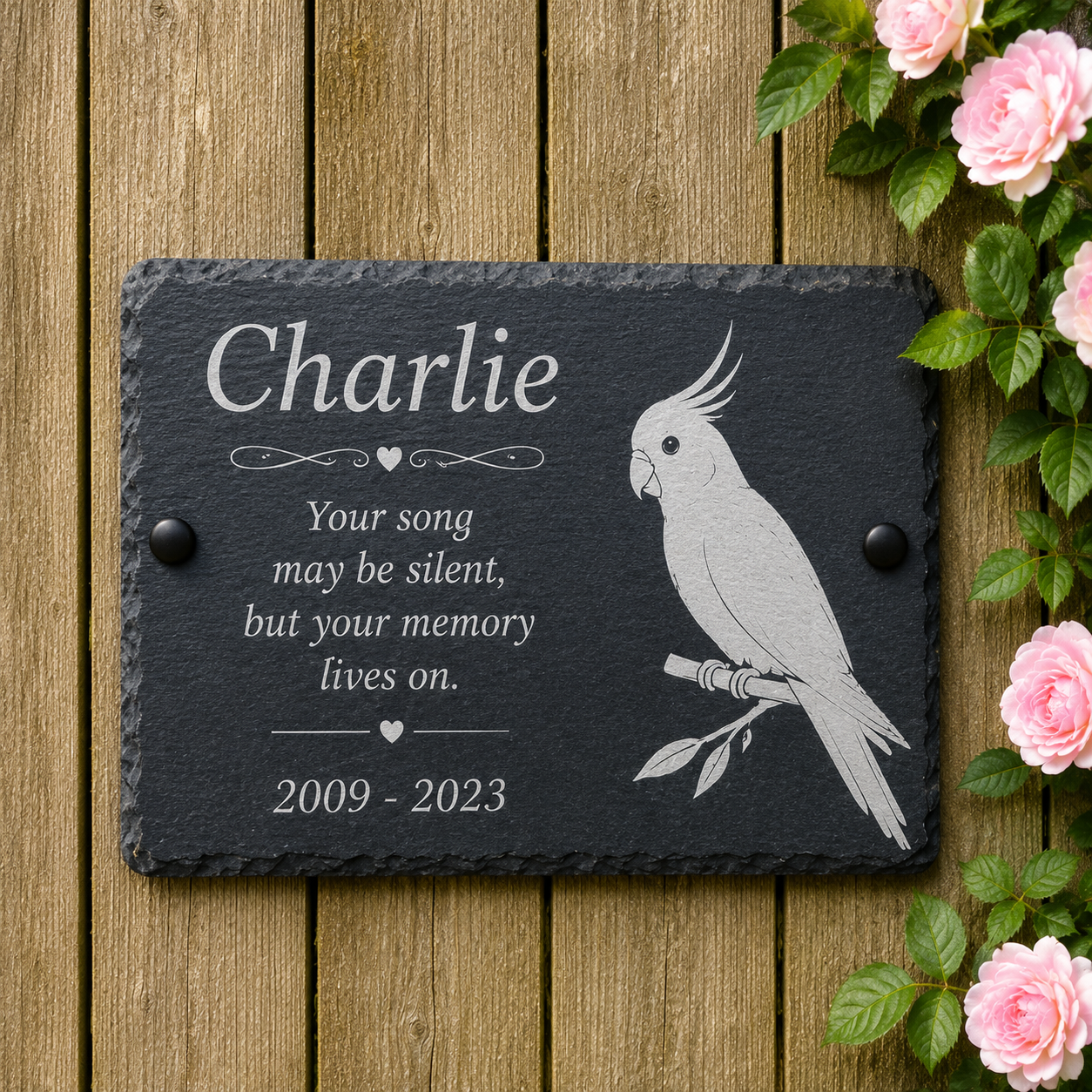 Personalized dark slate memorial plaque with engraved text reading 'Charlie' and a white cockatoo illustration, placed on a wooden surface with pink roses along the right edge.