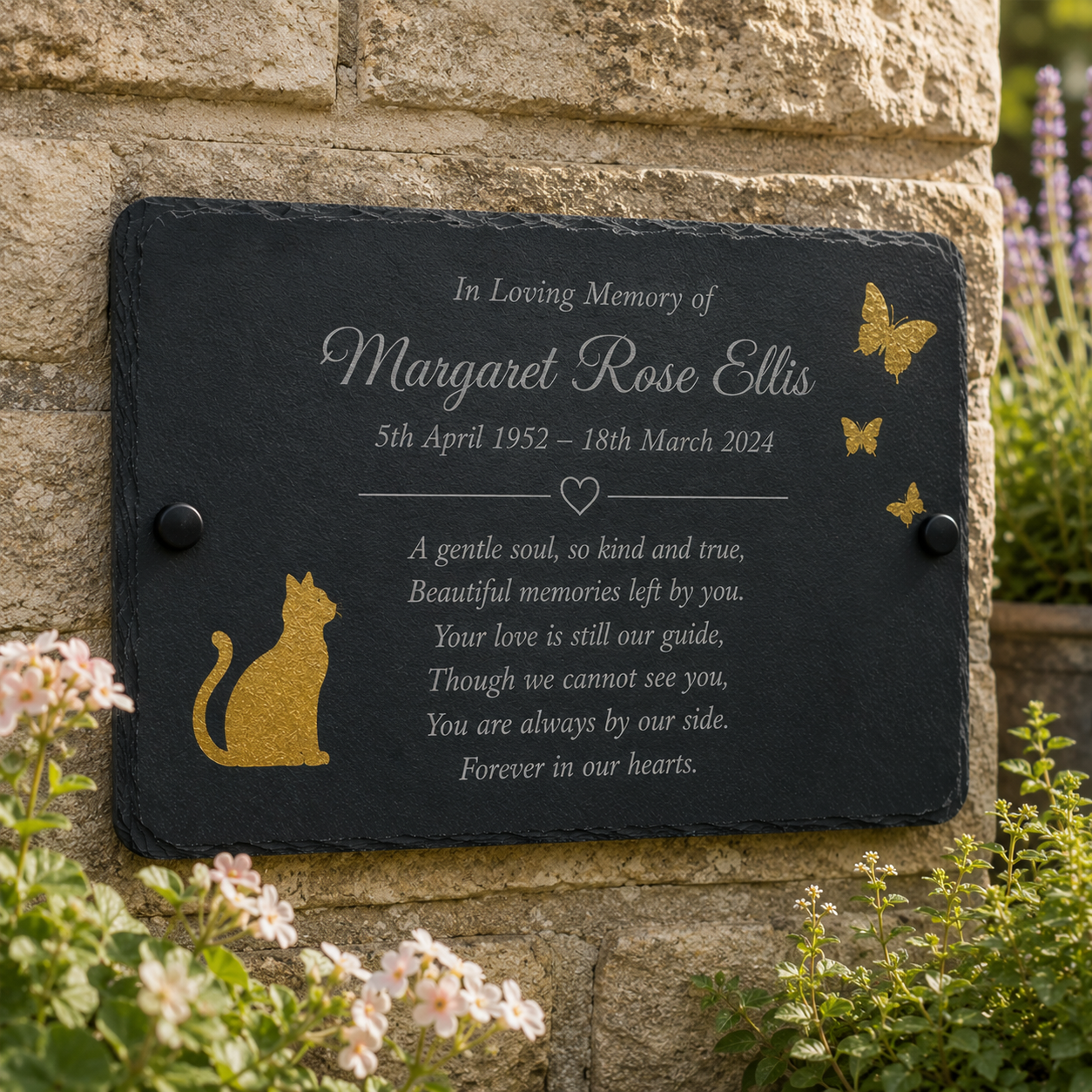 Personalized black slate memorial plaque with gold foil accents, mounted on a beige stone wall with flowers in the foreground.