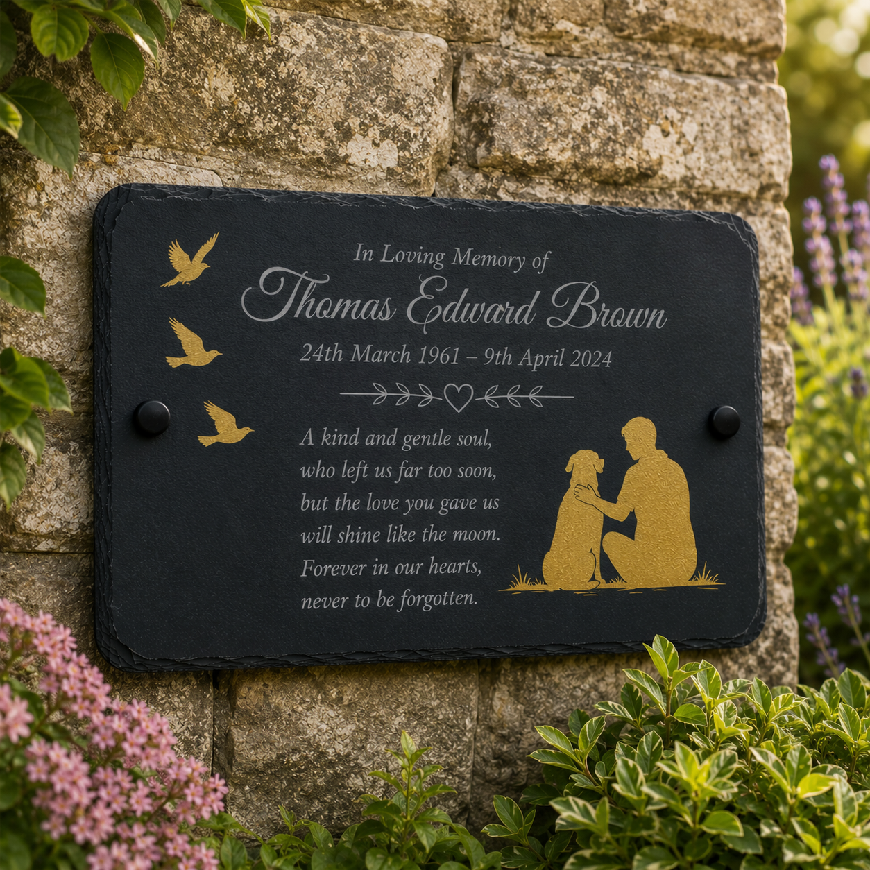 A personalized black slate memorial plaque with gold engraved silhouettes of birds, a seated figure, and a dog, mounted on a stone wall with greenery and flowers around it.