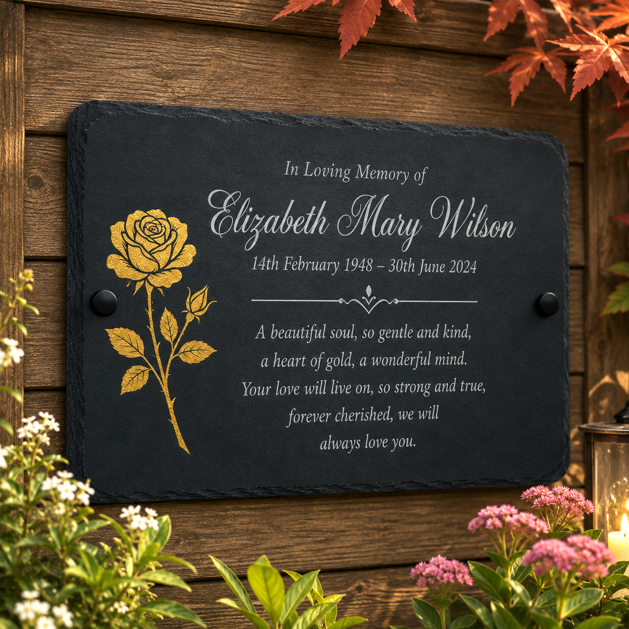 Personalized black slate memorial plaque with a gold rose illustration and white engraved text, mounted on a wooden wall with flowers and autumn leaves around it.