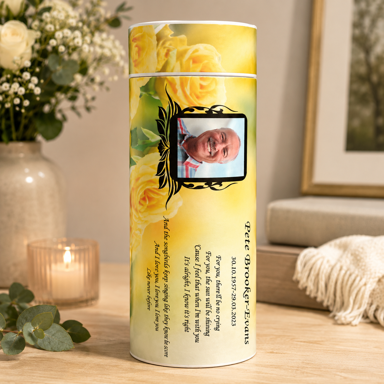 A personalized yellow rose memorial keepsake urn in a cylindrical container, displayed on a wooden table in a cozy, softly lit room with a lit candle, eucalyptus leaves, and a vase of flowers in the background.