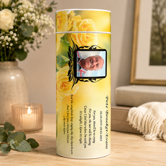 A personalized yellow rose memorial keepsake urn in a cylindrical container, displayed on a wooden table in a cozy, softly lit room with a lit candle, eucalyptus leaves, and a vase of flowers in the background.