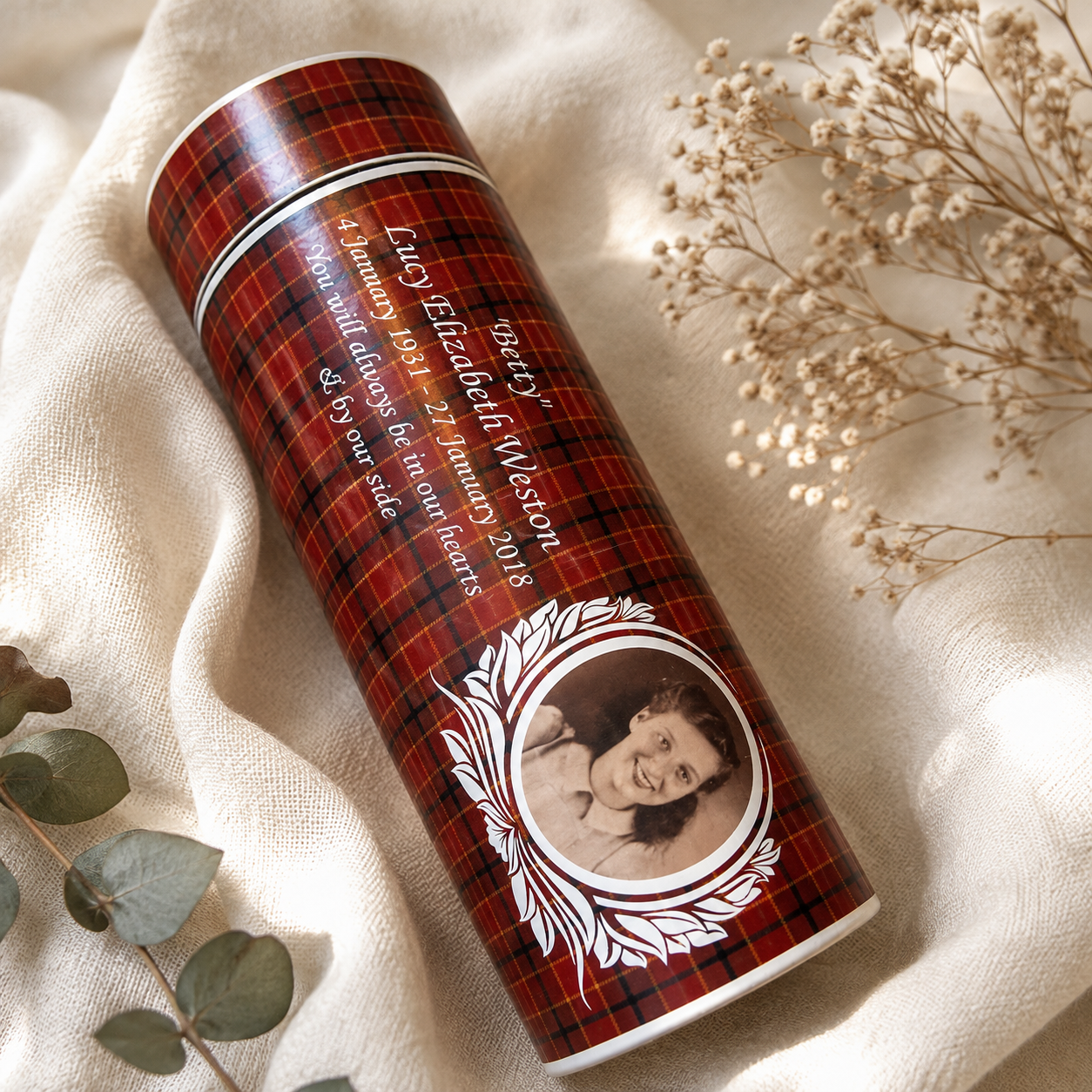 A personalized cylindrical tumbler with a red and black plaid pattern, featuring a printed portrait of a woman and white text, styled on a beige fabric background with dried flowers and green leaves.